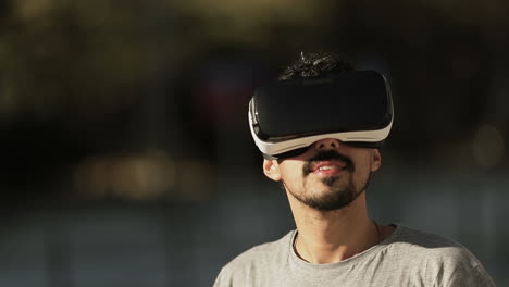 Close-up-shot-of-young-Arabic-mans-face-with-dark-curly-hair-and-beard-in-grey-T-shirt-being-in-park-in-virtual-reality-glasses